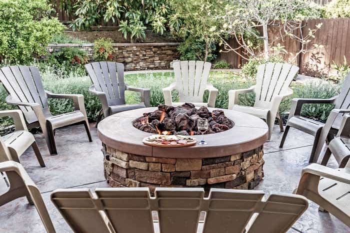 Large outdoor fire pit surrounded by wooden rocking chairs, beautifully landscaped backyard, with the glass of wine and food platter