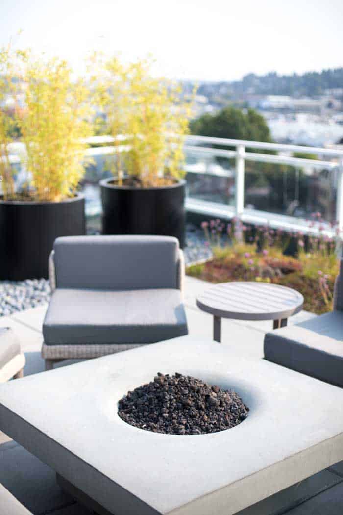 Shallow depth of field close up of square concrete gas fire pit with crushed pumice lava rock center in modern lifestyle condominium rooftop sitting area scene with padded cushion lounge chairs