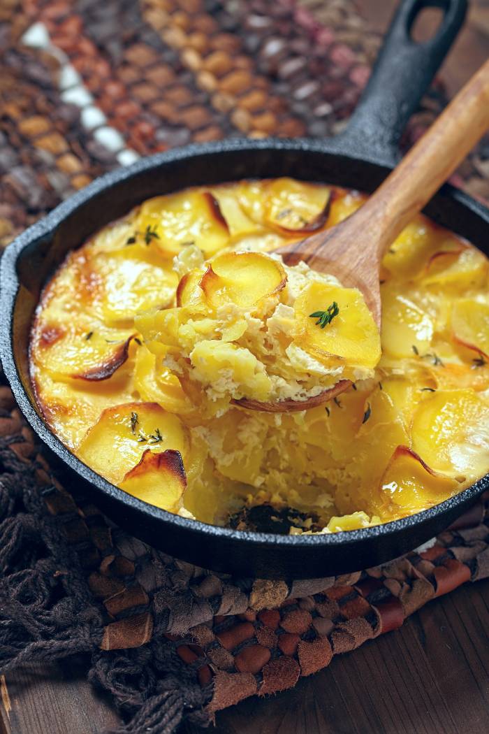 wooden spoon serving scalloped potatoes cooked in a cast iron skillet over grill fire